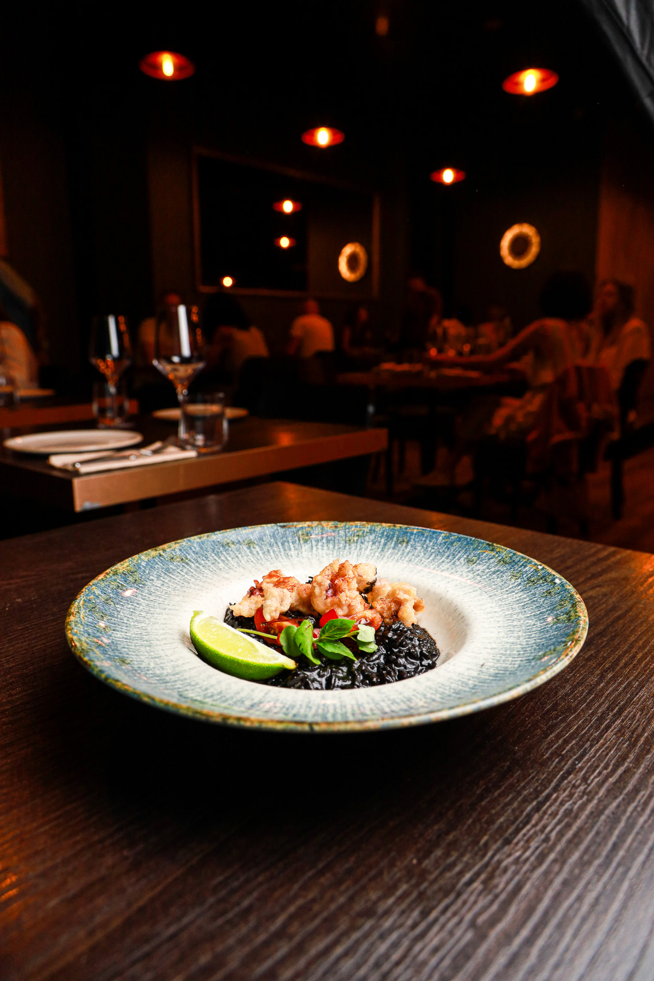 Black rice with fried squid served on a blue ceramic plate, with the softly lit Enora dining room behind
